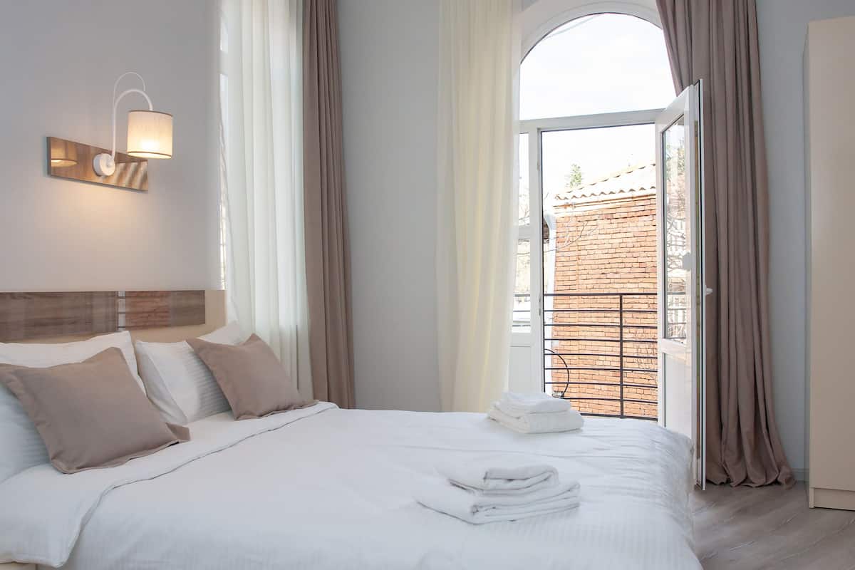 A hotel room with a bed, a wall-mounted light fixture, and a view of a brick building through an open door.