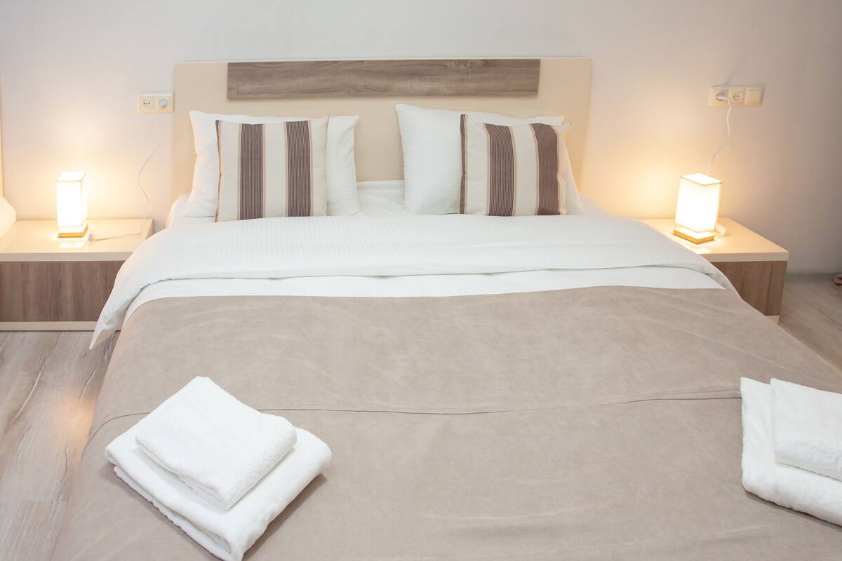 A neatly made bed with white linens and pillows, two bedside lamps, and folded towels on the floor.
