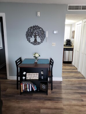 Un comedor con una mesa, sillas, un cuadro de pared con motivos de árbol, un florero con flores y una estantería con libros.