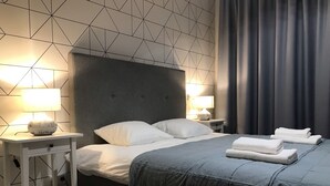 A neatly made bed with white linens and a grey headboard, flanked by bedside tables with lamps and a small shelf. - Crystal Apartments (Warsaw)