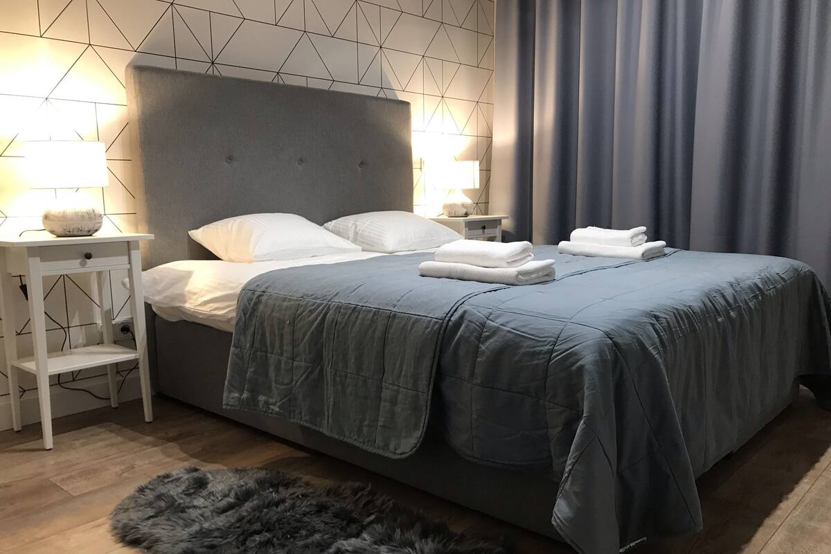 A modern bedroom with a grey upholstered headboard, white bedding, and a grey furry rug on a wooden floor.