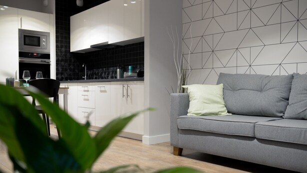 A modern kitchen with white cabinets, a black backsplash, and a grey sofa with a light green pillow. - Crystal Apartments (Warsaw)