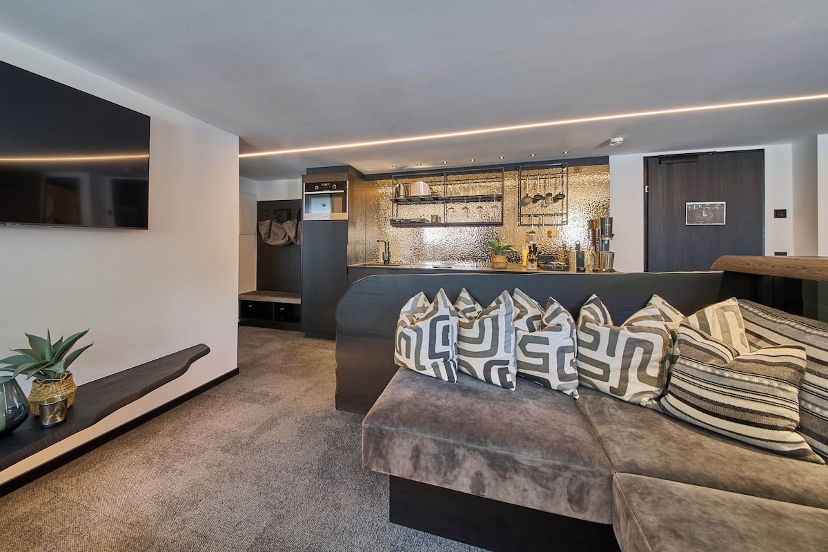 Uma sala moderna com um sofá cinza, uma TV de tela plana na parede e uma área de cozinha com bancada e armários.