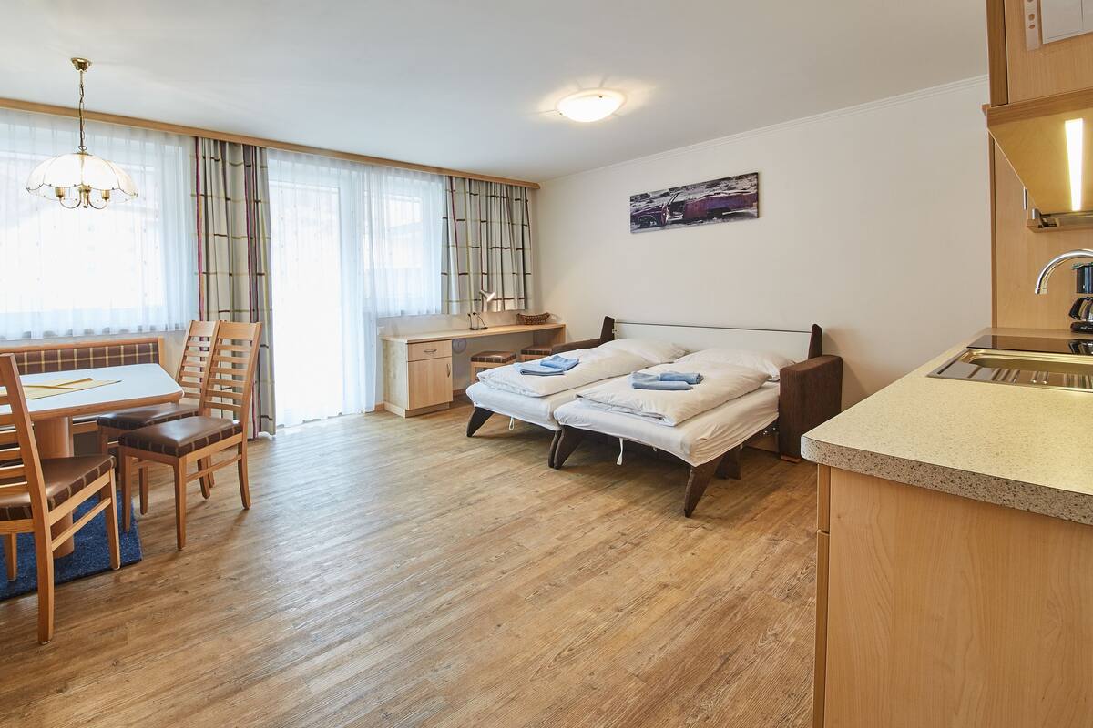 Quarto compacto com mesa de jantar, duas camas, pequena cozinha e janela com cortinas.