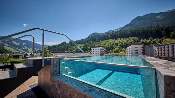 A modern outdoor swimming pool with clear water, surrounded by a wooden deck and stone walls, set against a backdrop of mountains and greenery. - ThePoolhouse Boutique Lodge (Saalbach-Hinterglemm)