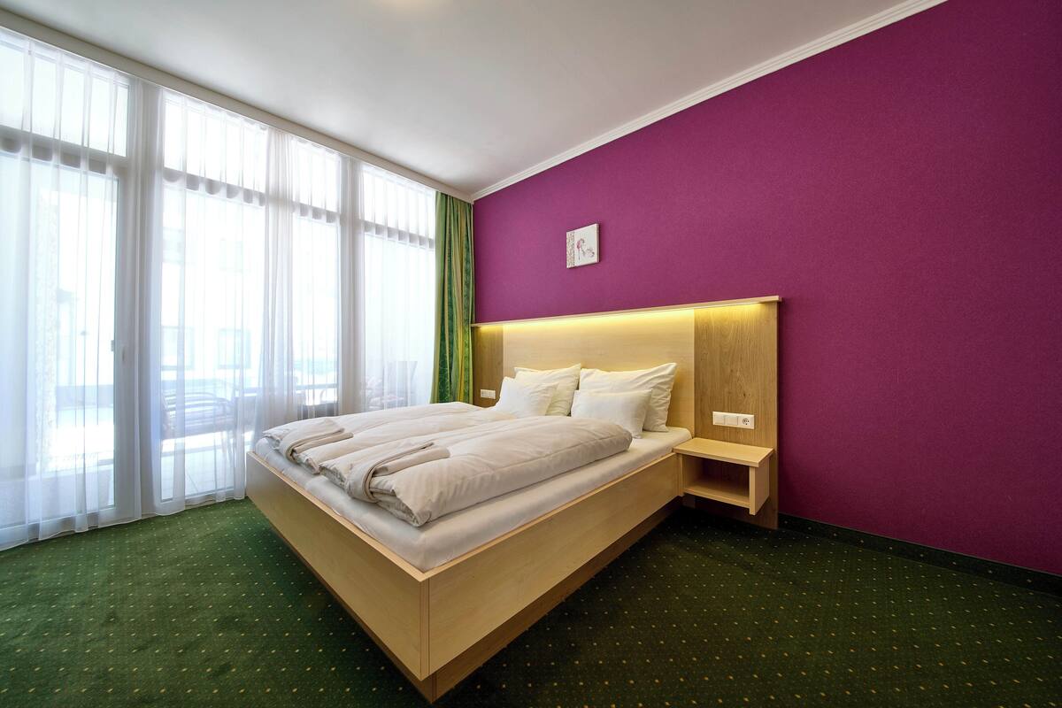 Quarto de hotel com uma cama grande, cabeceira de madeira, mesa de cabeceira e parede roxa.