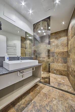 A modern bathroom with a large mirror, a white sink, and a shower area with stone tiles. - Appartement Bretterbude (Saalbach-Hinterglemm)