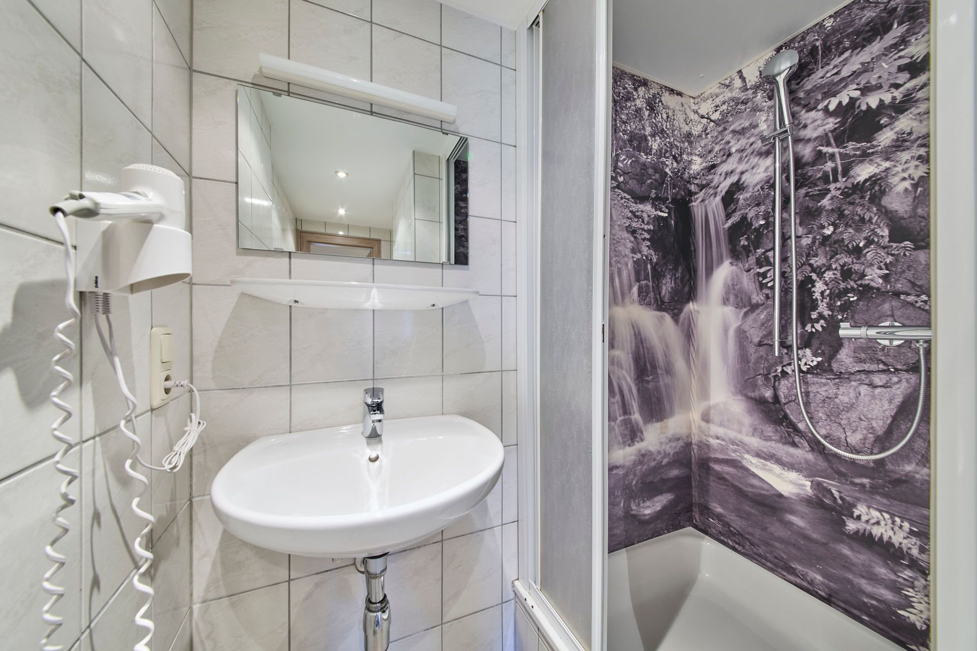 A bathroom with a white sink, a mirror, a hairdryer, and a shower with a waterfall mural.