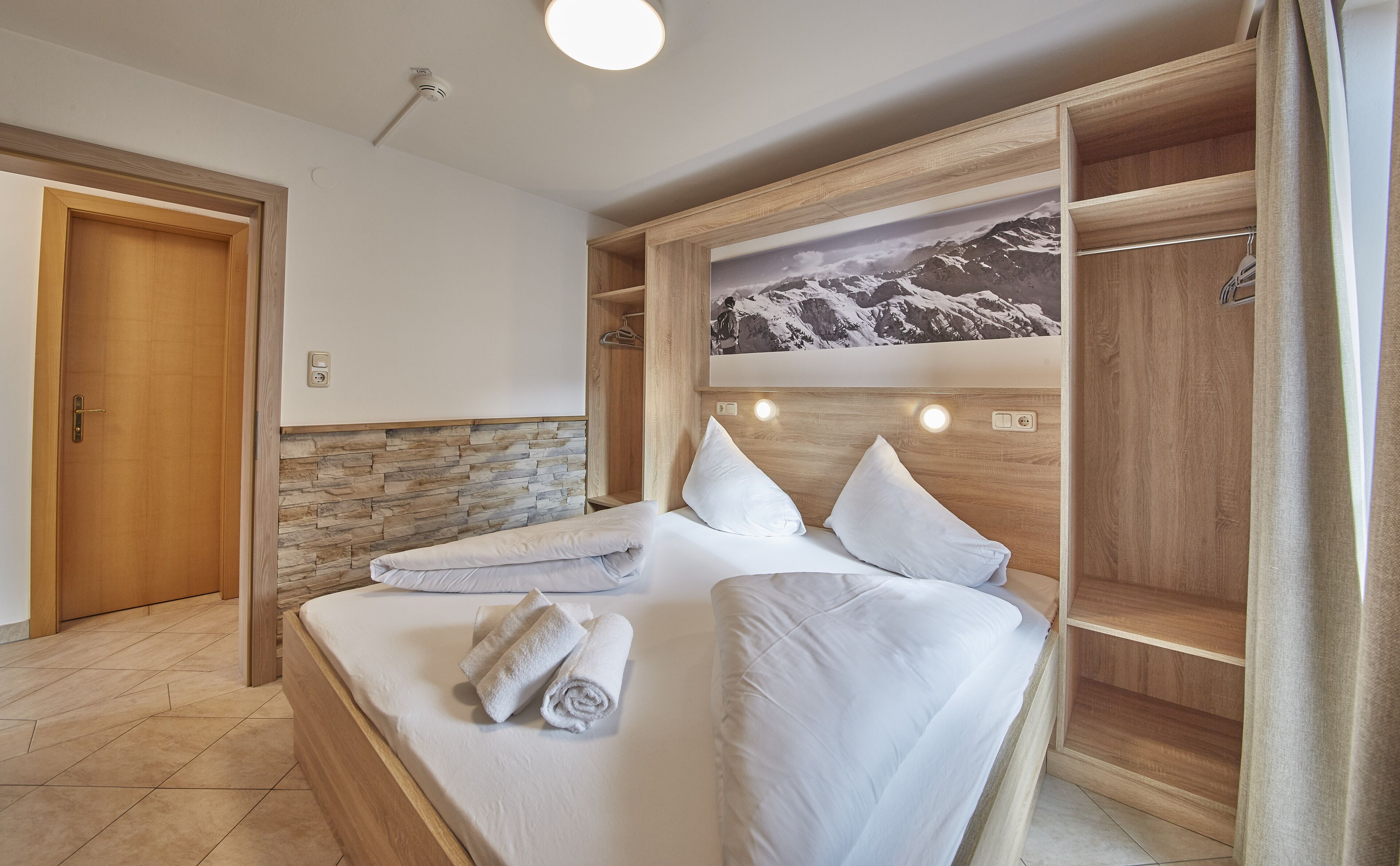 A bedroom with a wooden headboard, a bed with white linens, a stone accent wall, and a mountain painting above the bed.