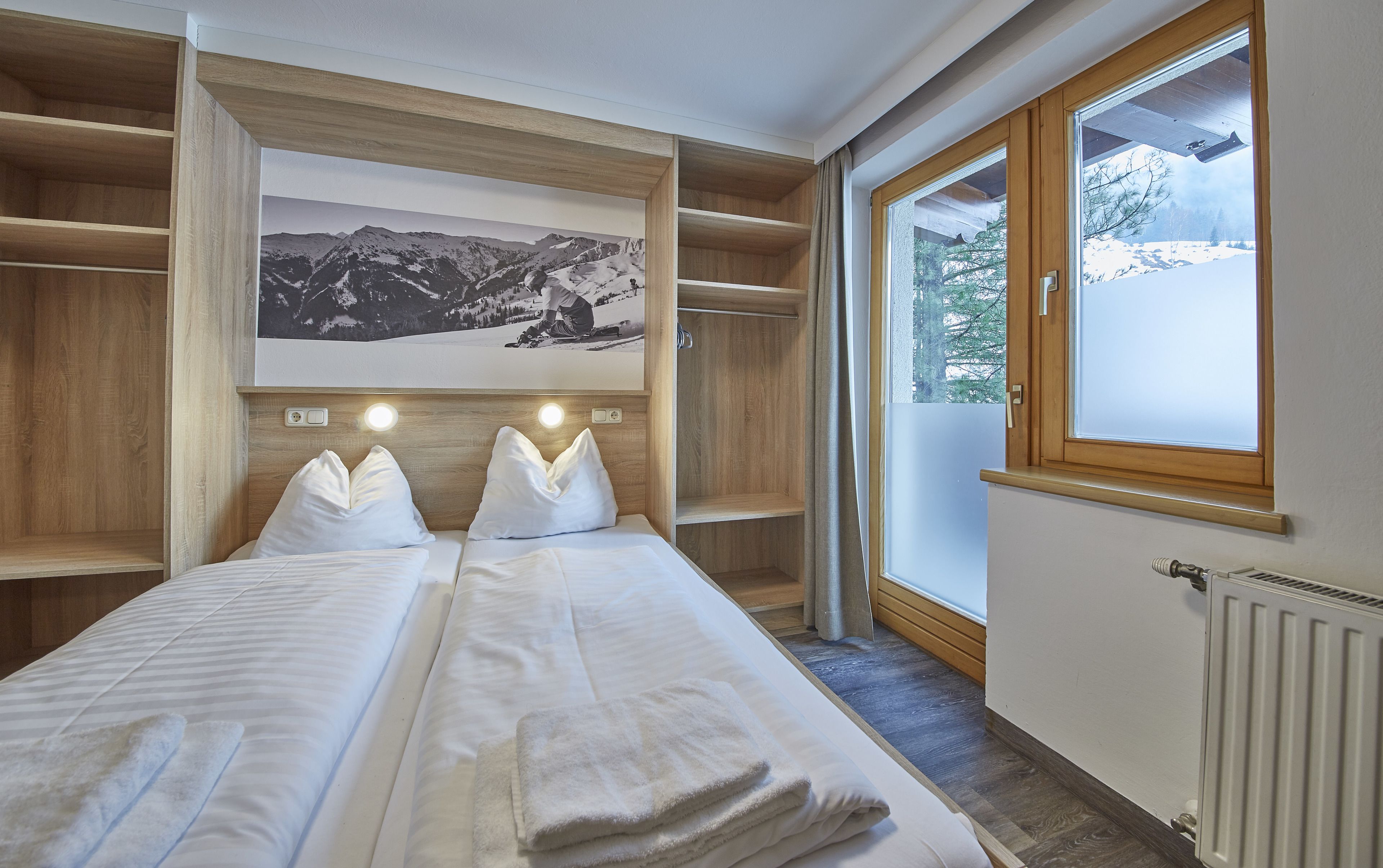 A hotel room with two beds, wooden shelving, a mountain view, and a radiator.