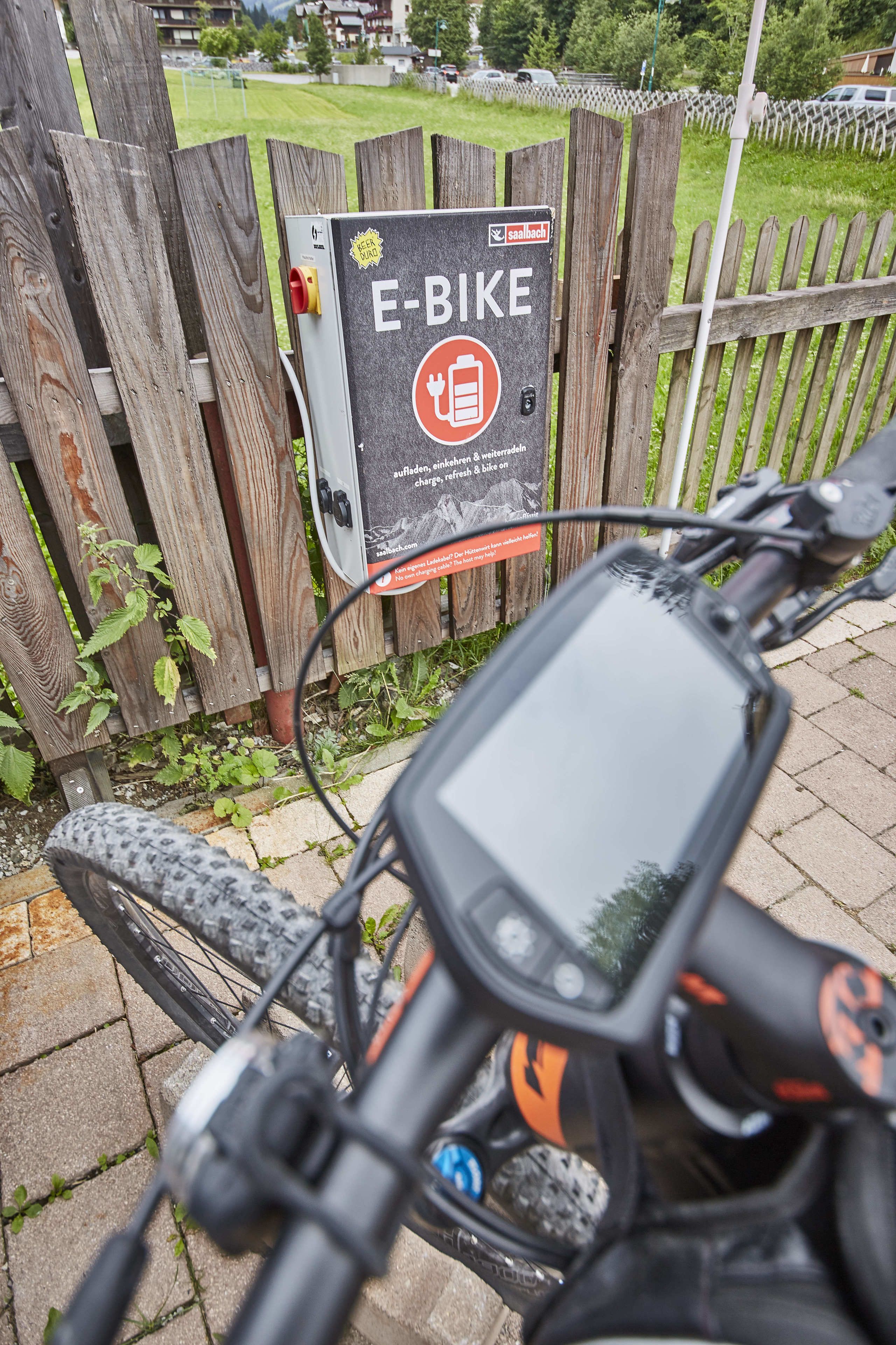 A close-up of a bicycle with a digital display and a charging station labeled "E-BIKE" in front of a wooden fence.