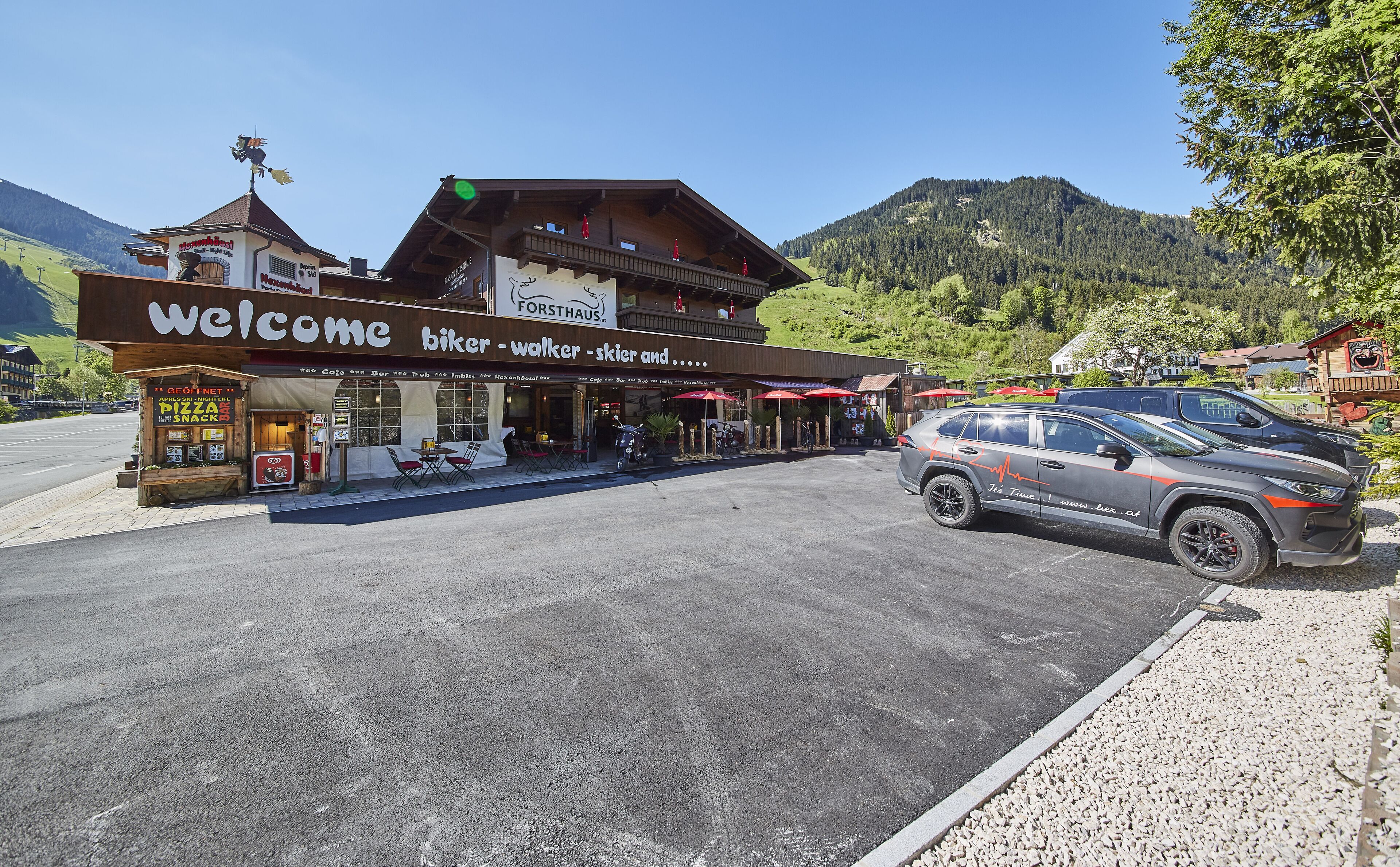 Un chalet de montagne avec une pancarte « Bienvenue », des sièges à l’extérieur et un VUS garé.