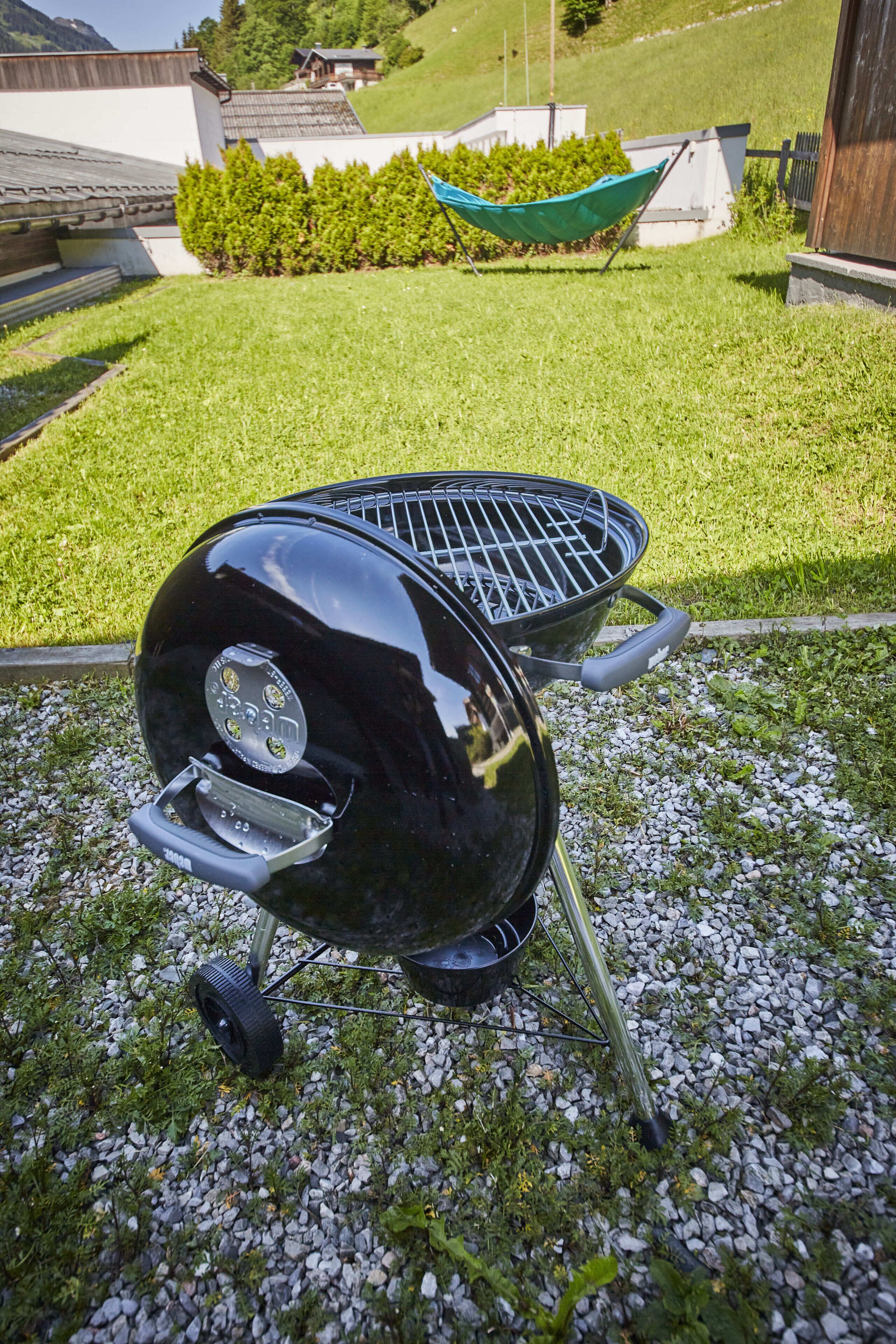 A black barbecue grill with a digital display, positioned on a gravel surface in a grassy area with a hammock and a building in the background.