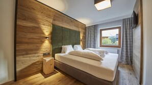 A modern bedroom with a wooden headboard, a large bed with white linens, a bedside table, and a window with curtains. - Appartements Gappmaier (Saalbach-Hinterglemm)