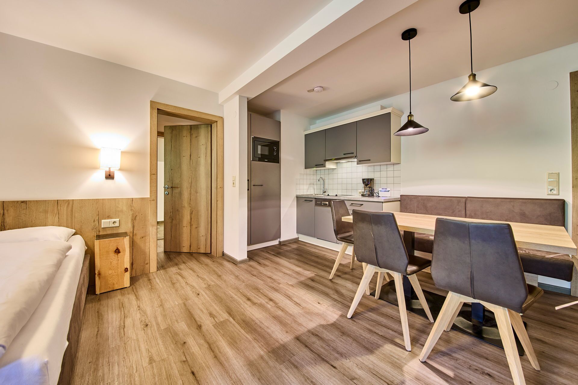 A modern hotel room with a wooden floor, a bed, a wooden nightstand, a desk, and a dining area with chairs and a bench.