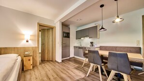 A modern hotel room with a wooden floor, a bed, a wooden nightstand, a desk, and a dining area with chairs and a bench. - Appartements Gappmaier (Saalbach-Hinterglemm)