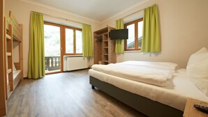A bedroom with a bed, a TV mounted on the wall, a wooden wardrobe, and a window with curtains. - Holiday Lodge Central (Saalbach-Hinterglemm)