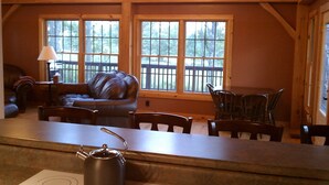 Ein gemütliches Wohnzimmer mit Ledersofa, Holzstühlen, einem Couchtisch und großen Fenstern mit Blick auf Bäume.