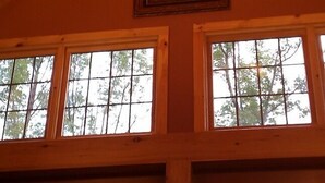 Ein Zimmer mit hoher Decke und Holzbalken, große Fenster mit Blick auf Bäume und eine kleine Leuchte an der Decke.