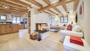 Amplia sala de estar con chimenea, vigas de madera, cocina con armarios de madera y sofá blanco con cojines rojos.