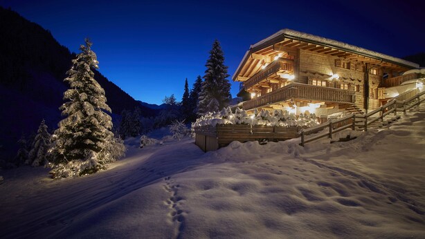 Un albergue de montaña cubierto de nieve, con fachada de madera y un balcón que da a un paisaje nevado.