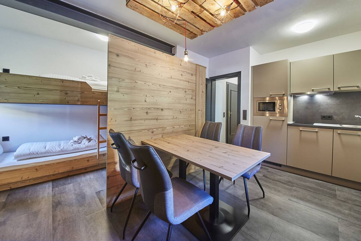 A compact living space with a bunk bed, a dining table with chairs, a kitchenette, and a wooden accent wall.