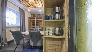 A cozy room with a wooden cabinet containing a coffee maker and mugs, a dining table with chairs, and a window with curtains. - Time for Sport Appartements (Saalbach-Hinterglemm)