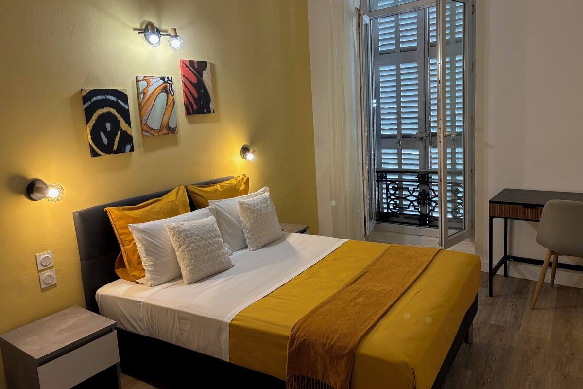 A hotel room with a bed, bedside table, wall art, and a window with blinds.