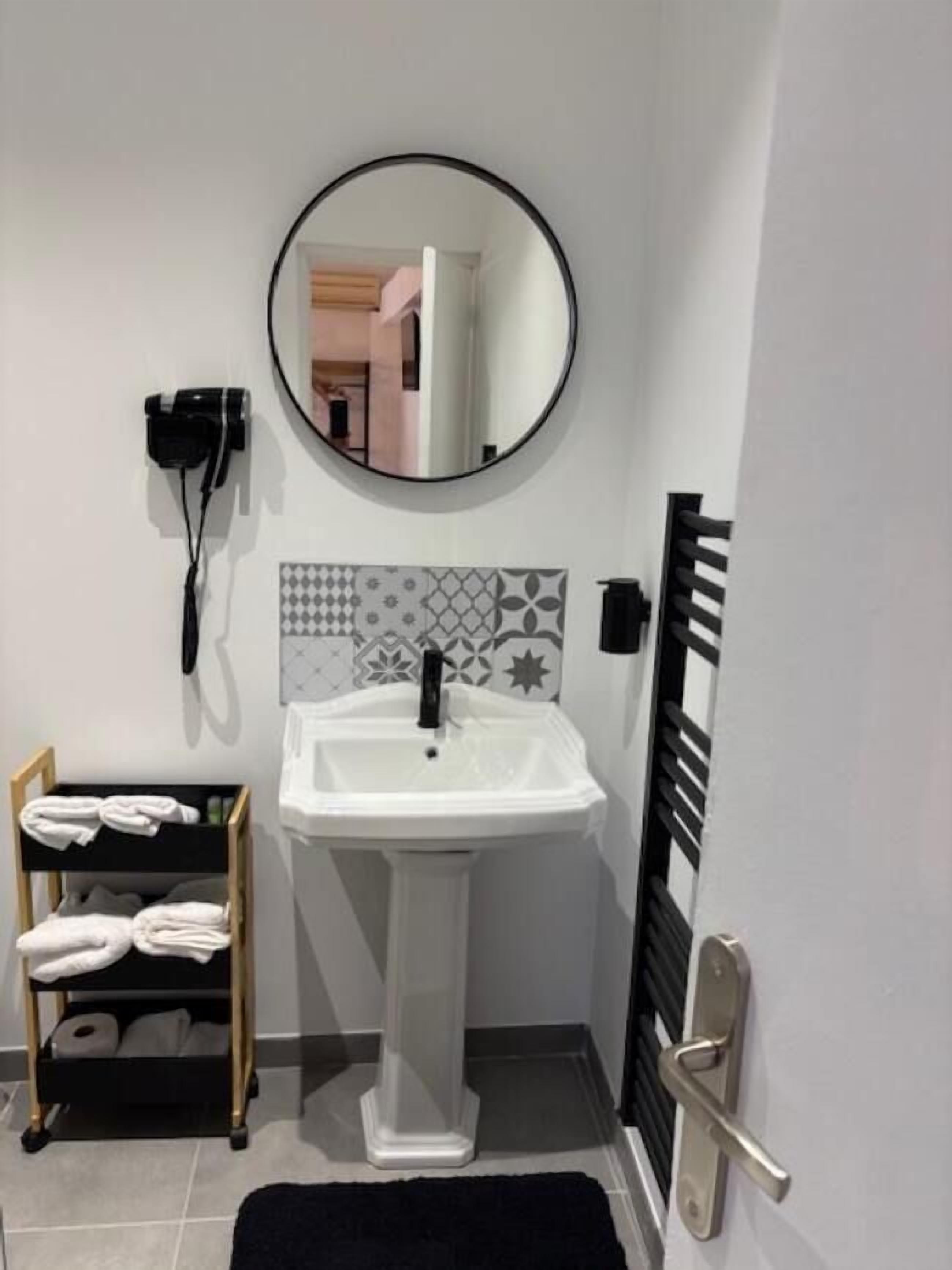 A bathroom with a white pedestal sink, a round mirror, a towel rack, and a black towel shelf.