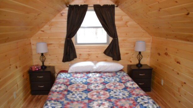 Ein Schlafzimmer mit Holzbalkendecke, einem Bett mit floraler Steppdecke, zwei Nachttischen mit Lampen und einem Fenster mit Vorhängen.
