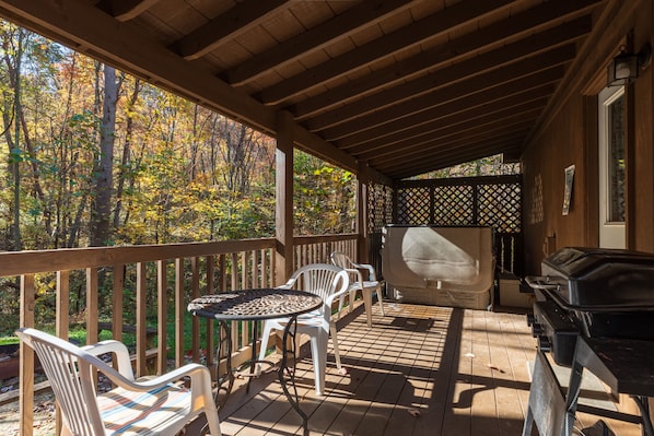 A covered outdoor deck with a grill, chairs, and a view of autumn trees.