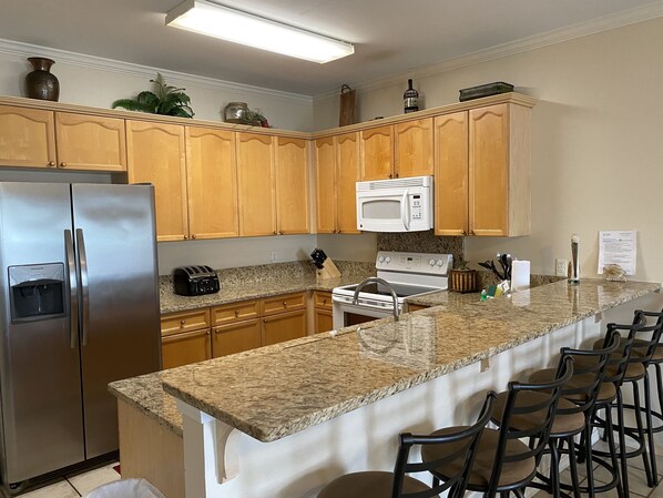 A kitchen with wooden cabinets, a stainless steel refrigerator, a microwave, a stove, and a granite countertop. There are bar stools along the counter.