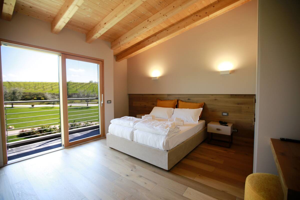 A bedroom with a large bed, wooden ceiling beams, a nightstand, and a view of a vineyard through a sliding door.