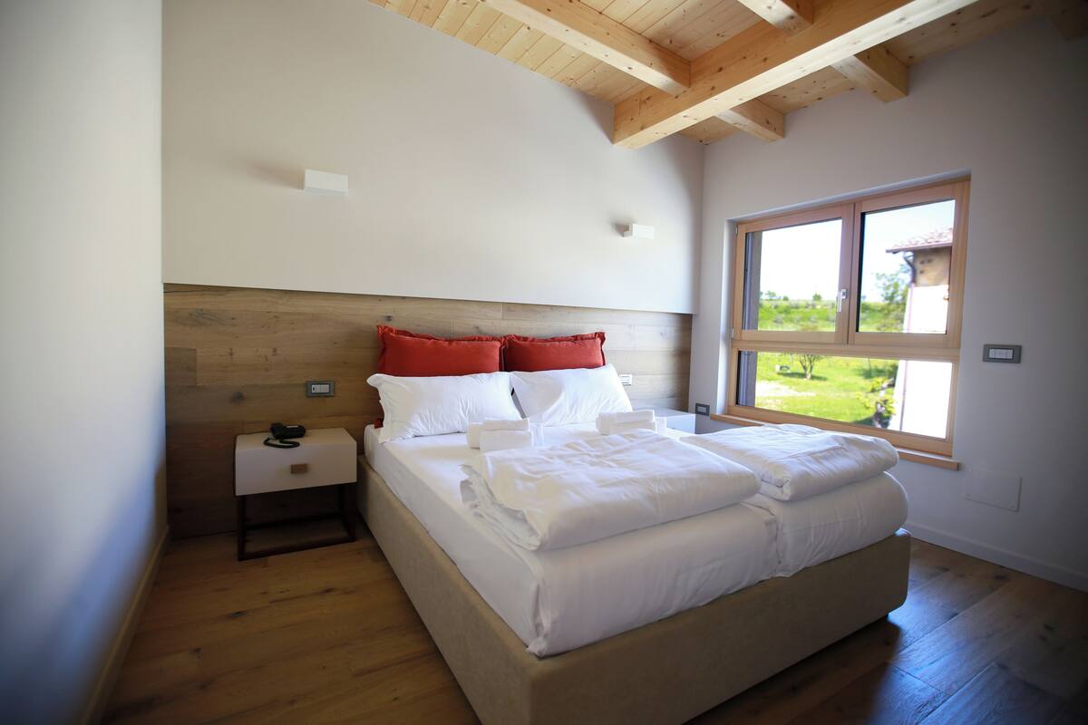A bedroom with a wooden ceiling, a large window, a bed with white bedding, and a nightstand with a phone.