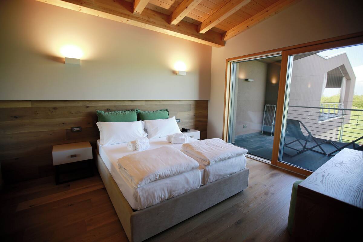 A modern bedroom with a wooden headboard, a large bed with white linens, two wall-mounted lights, and a sliding glass door leading to an outdoor area.