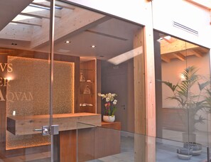 Un área de recepción con mostrador de madera, divisiones de vidrio y una planta en maceta con flores blancas.