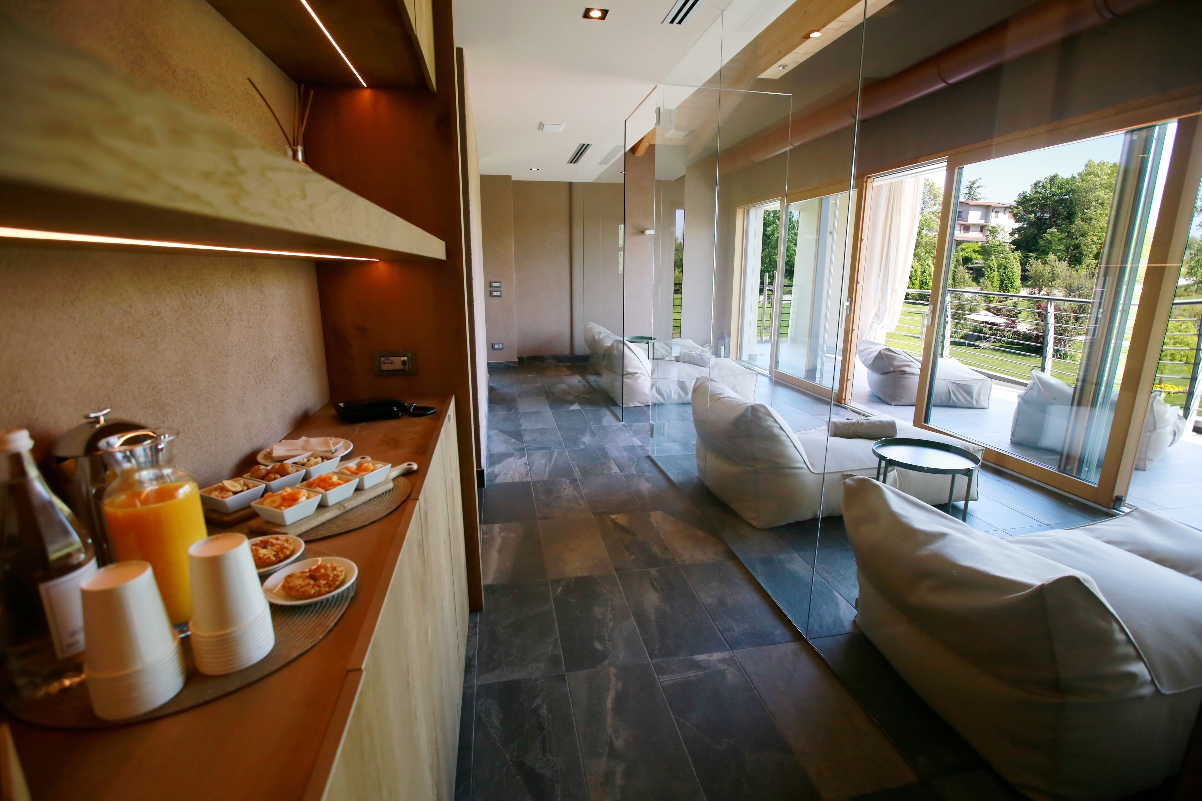 A modern hotel room with a breakfast buffet, a seating area with sofas, and a large window offering a view of the outdoors.