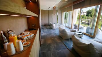 A modern hotel room with a breakfast buffet, a seating area with sofas, and a large window offering a view of the outdoors.