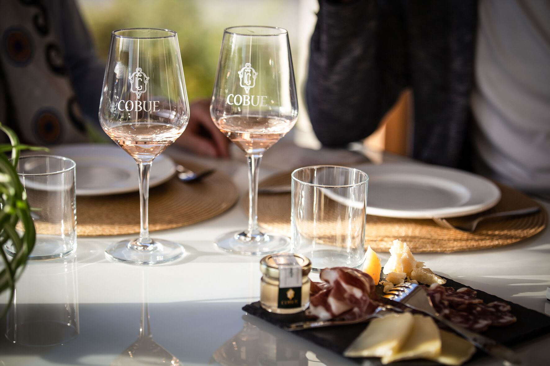 Una mesa dispuesta con copas de vino con la etiqueta 'COBUE', una botella pequeña y un plato con queso y embutidos.