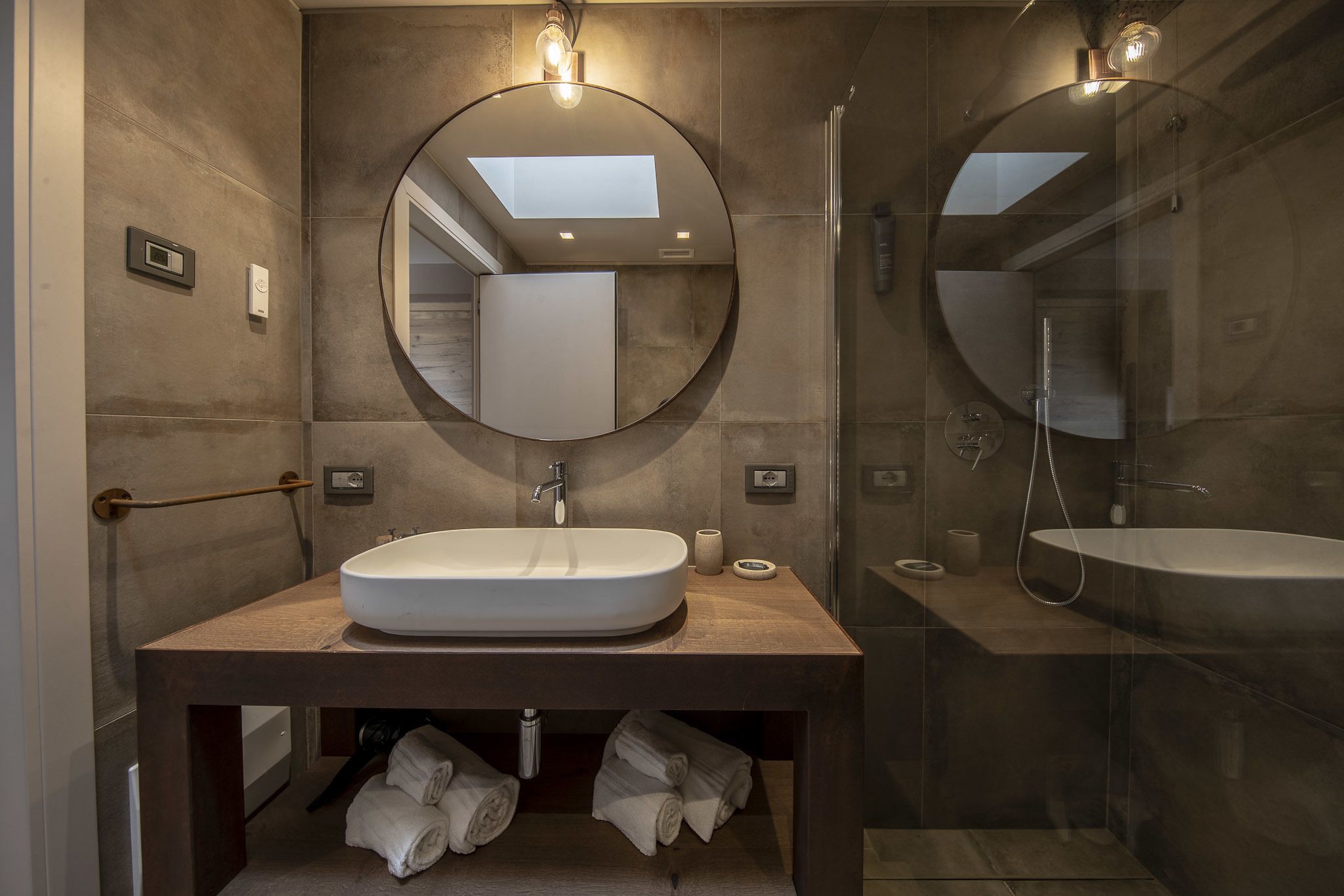 Een moderne badkamer met een groot ovaal wastafel, een ronde spiegel en een douche met glazen wanden.