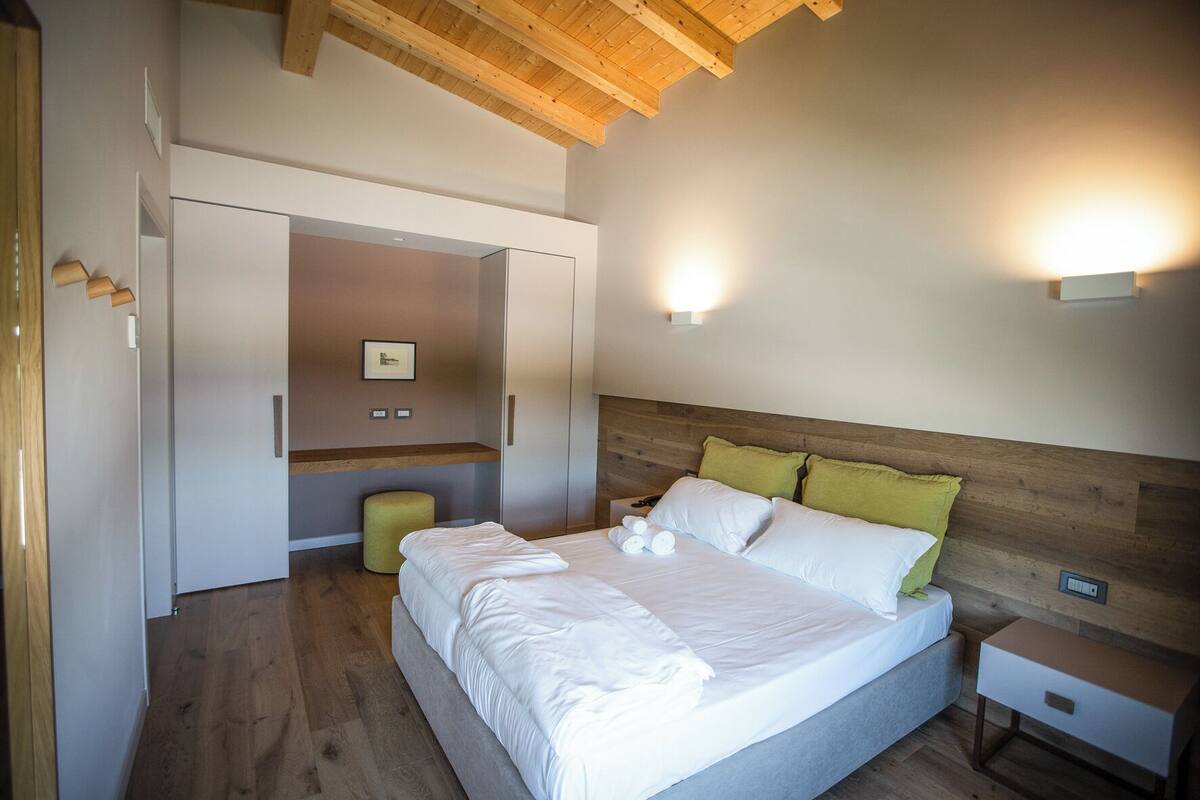 A bedroom with a wooden ceiling, a bed with white bedding and green pillows, a bedside table, and a small stool.