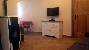Une pièce avec un téléviseur posé sur un meuble blanc, un canapé, une porte en bois et un sol carrelé.