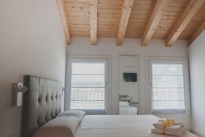 A bedroom with a large bed, wooden ceiling beams, two windows with blinds, a bedside table, and a chair. - Terra Ferma Rooms & More (Salzano)