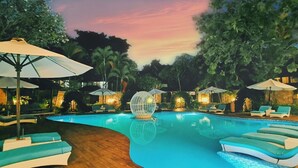 Un área de piscina con sillas de descanso y sombrillas, rodeada de árboles y un cielo al atardecer.