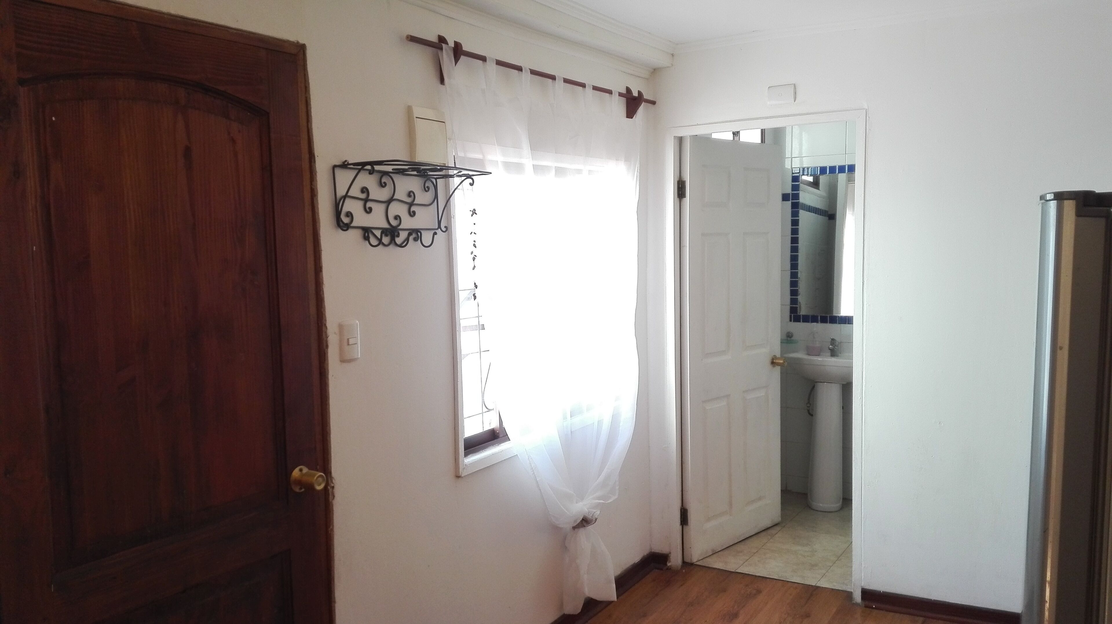 A hallway with a wooden door, a white wall, a window with sheer curtains, and a bathroom visible through an open door.