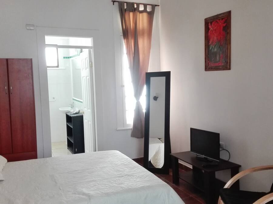 Un dormitorio con cama, armario, televisor, espejo y un baño visible a través de una puerta abierta.