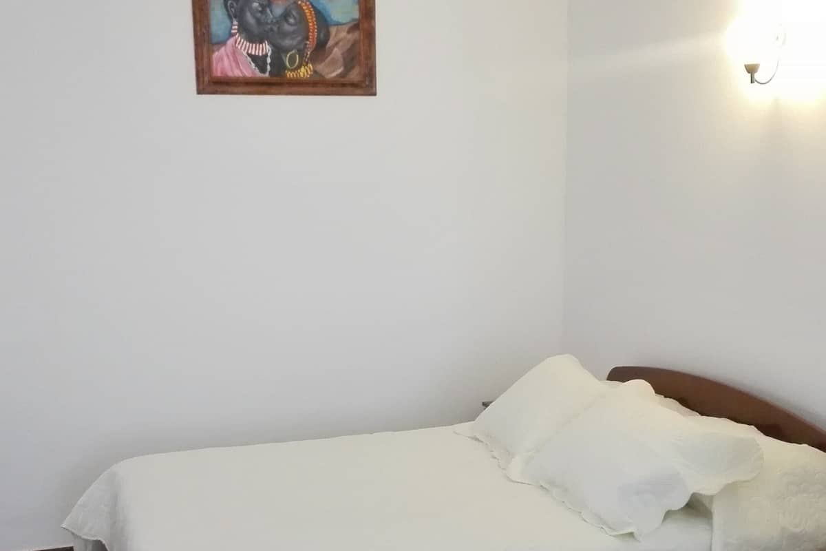Una cama pequeña y bien hecha con una colcha blanca en una habitación con suelo de baldosas y una única lámpara en la pared.