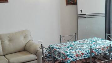 Una sala de estar con un sofá, una mesa de comedor con sillas, una lámpara de pared y un refrigerador.