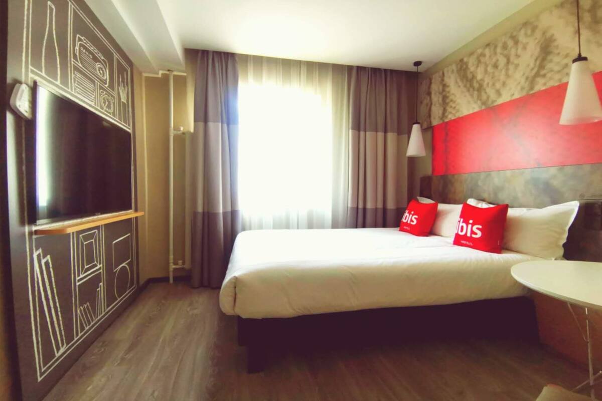 A hotel room with a large bed, a flat-screen TV, a small table, and a red accent wall.