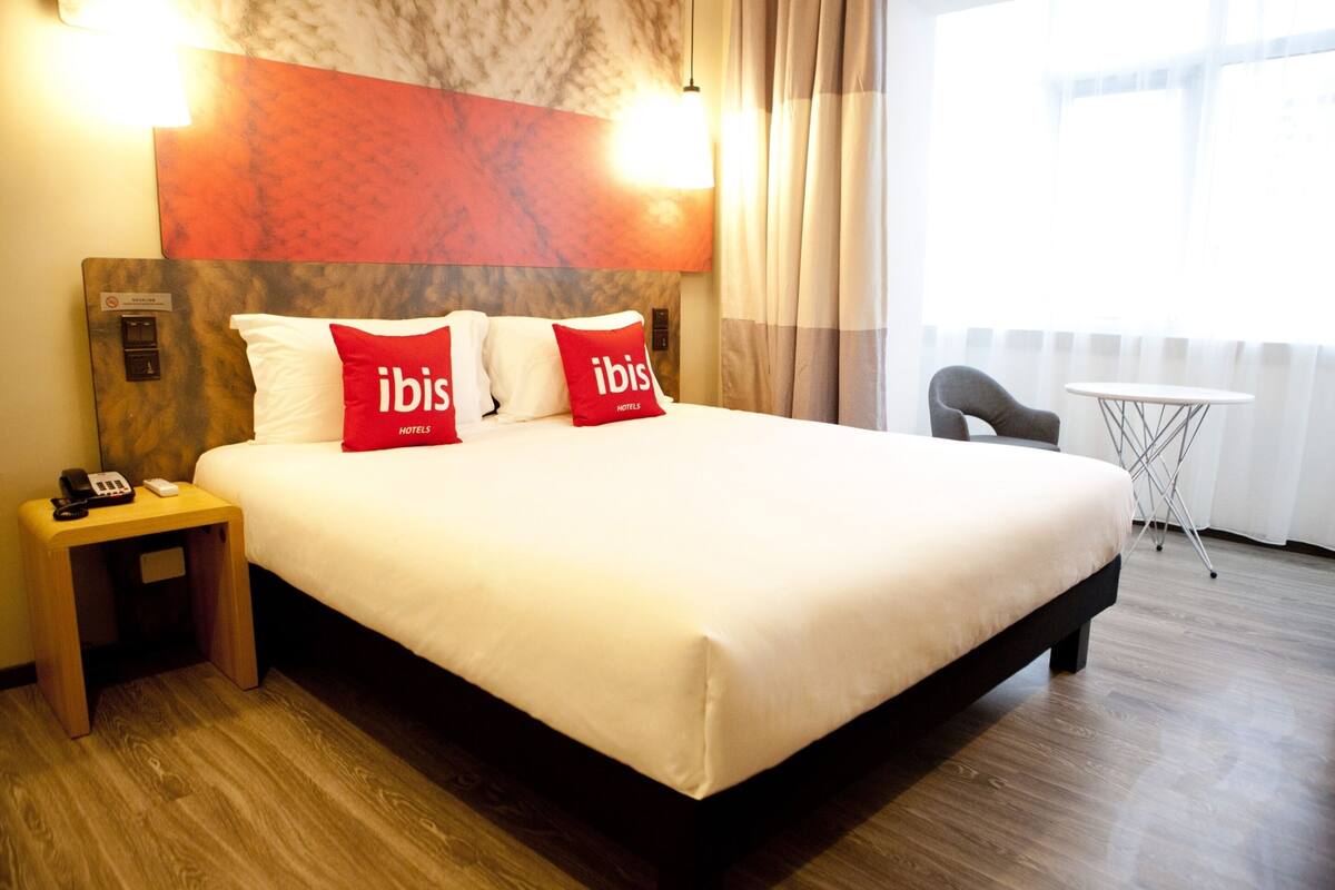 A hotel room with a large bed, two red pillows with "ibis Hotels" branding, a bedside table with a phone, and a small table with a chair near a window.